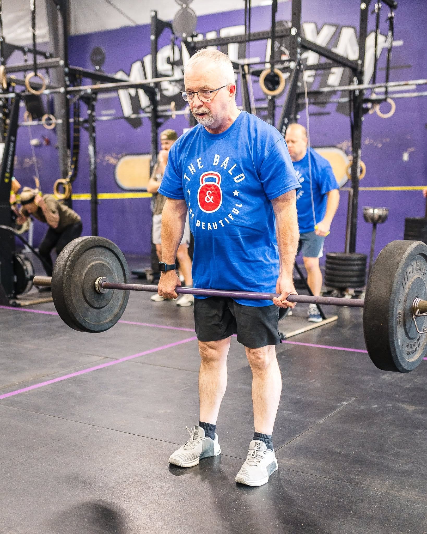 An older Misfit member standing over a loaded barbell under the gym's purple mural