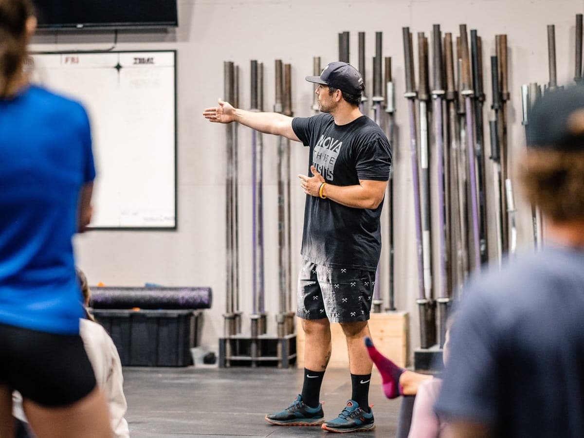A coach in a cap pointing across the floor as he briefs the seated class, barbells racked behind him