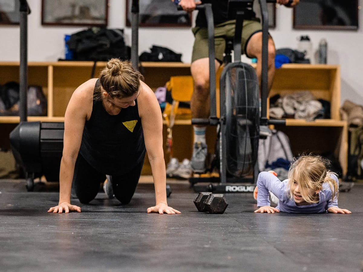 A member on hands and knees mid-workout grinning at a small girl doing the same movement beside her