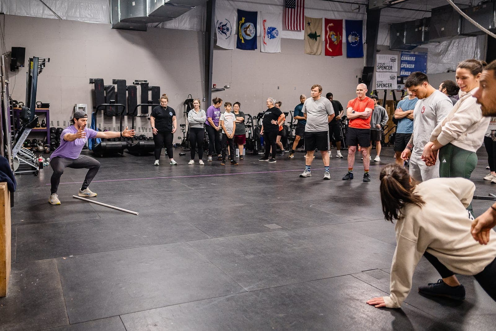 A coach demonstrating a squat with a PVC pipe in front of a half-circle of watching members