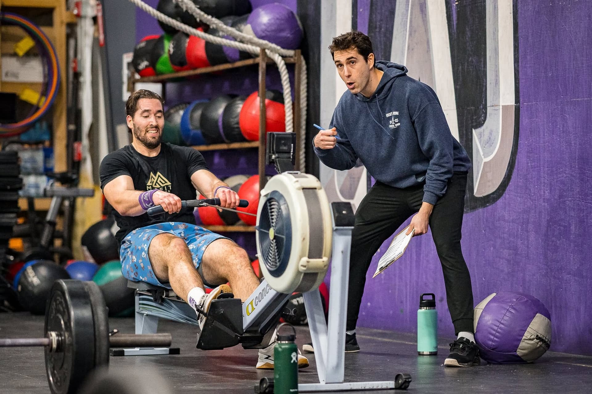 A coach with a clipboard leaning in to talk to a member mid-effort on the rower
