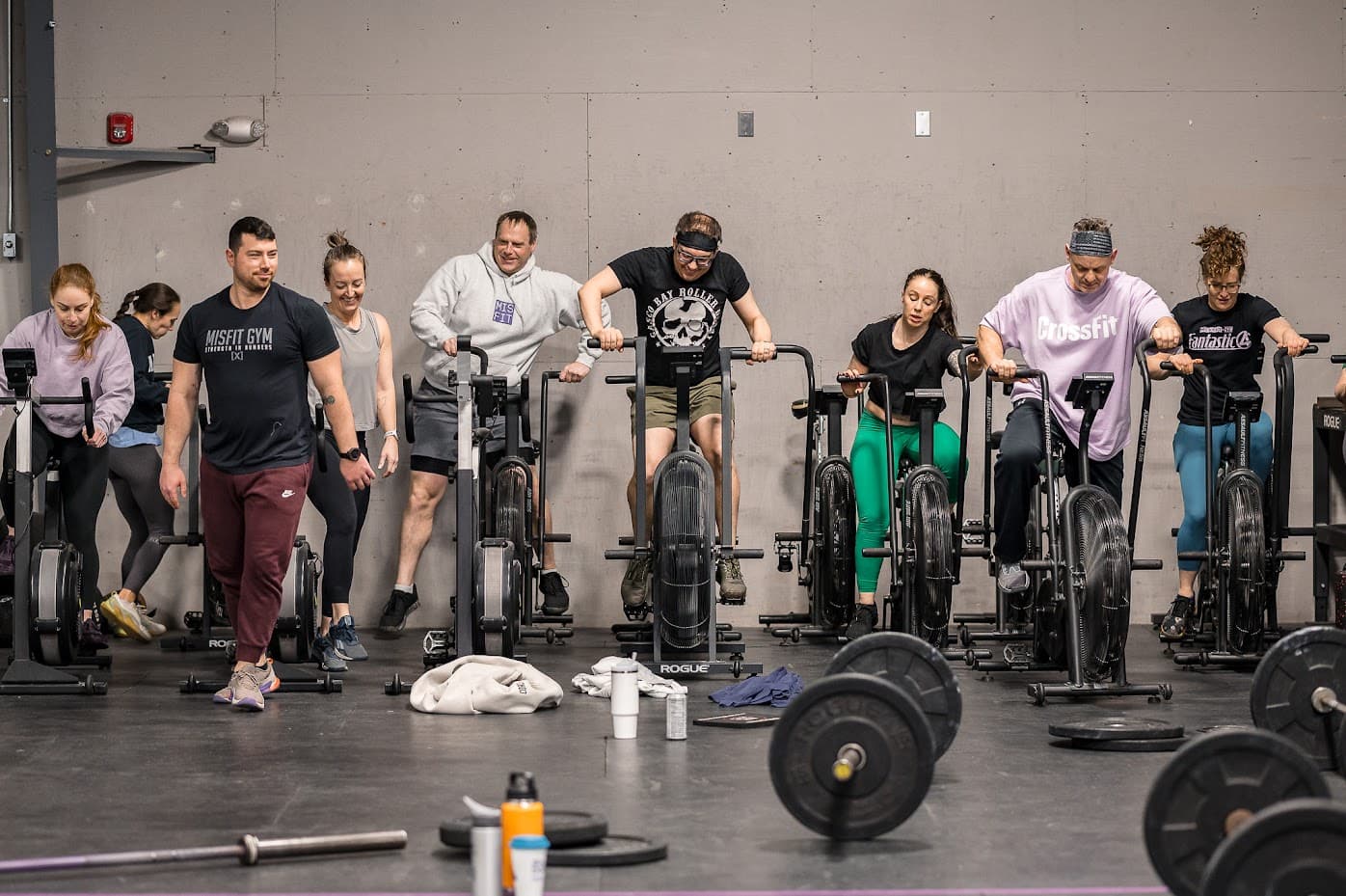 Nine members on air bikes in a row mid-workout, a coach walking the line in front of them