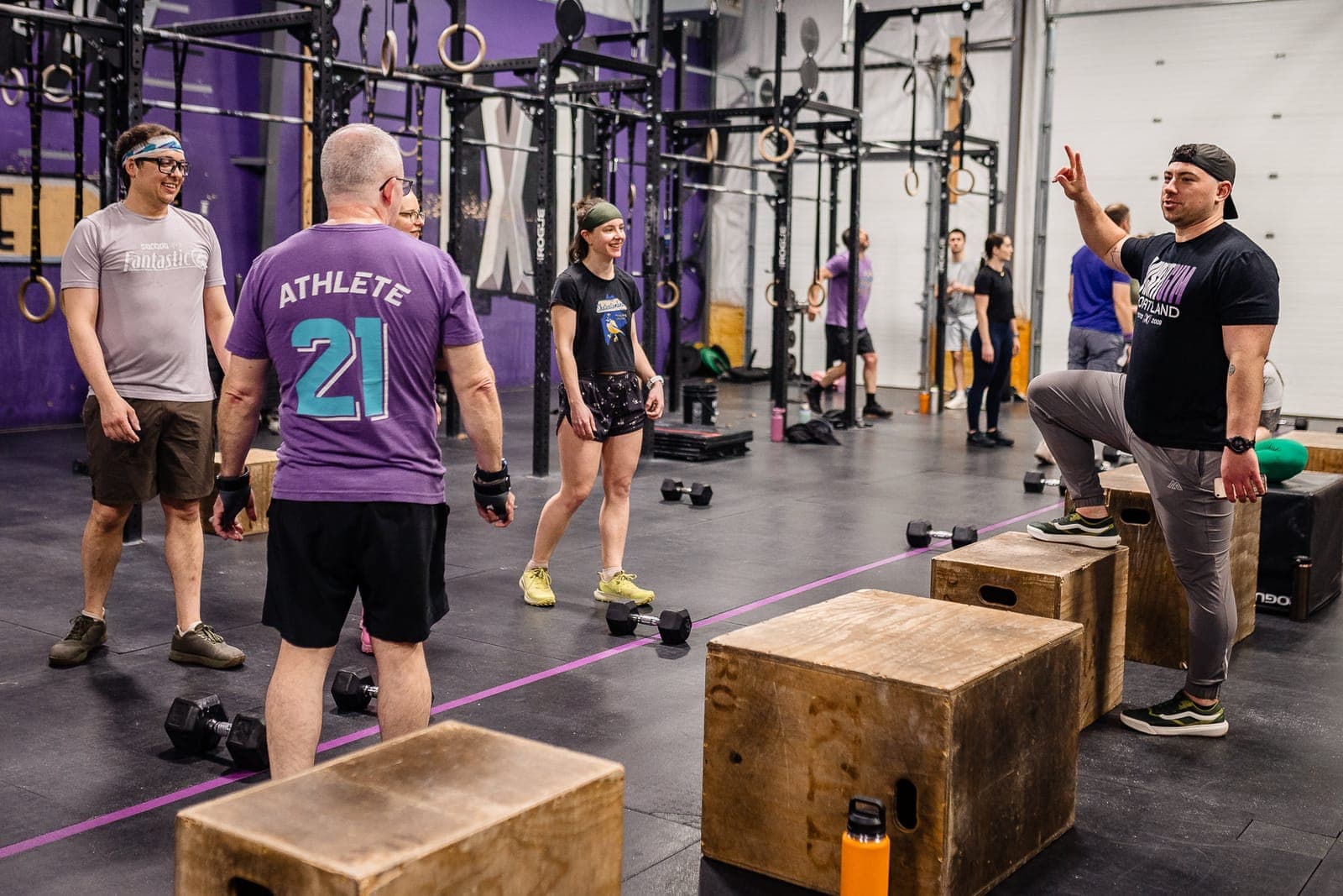 A coach with one foot up on a plyo box briefing a smiling class gathered under the rig